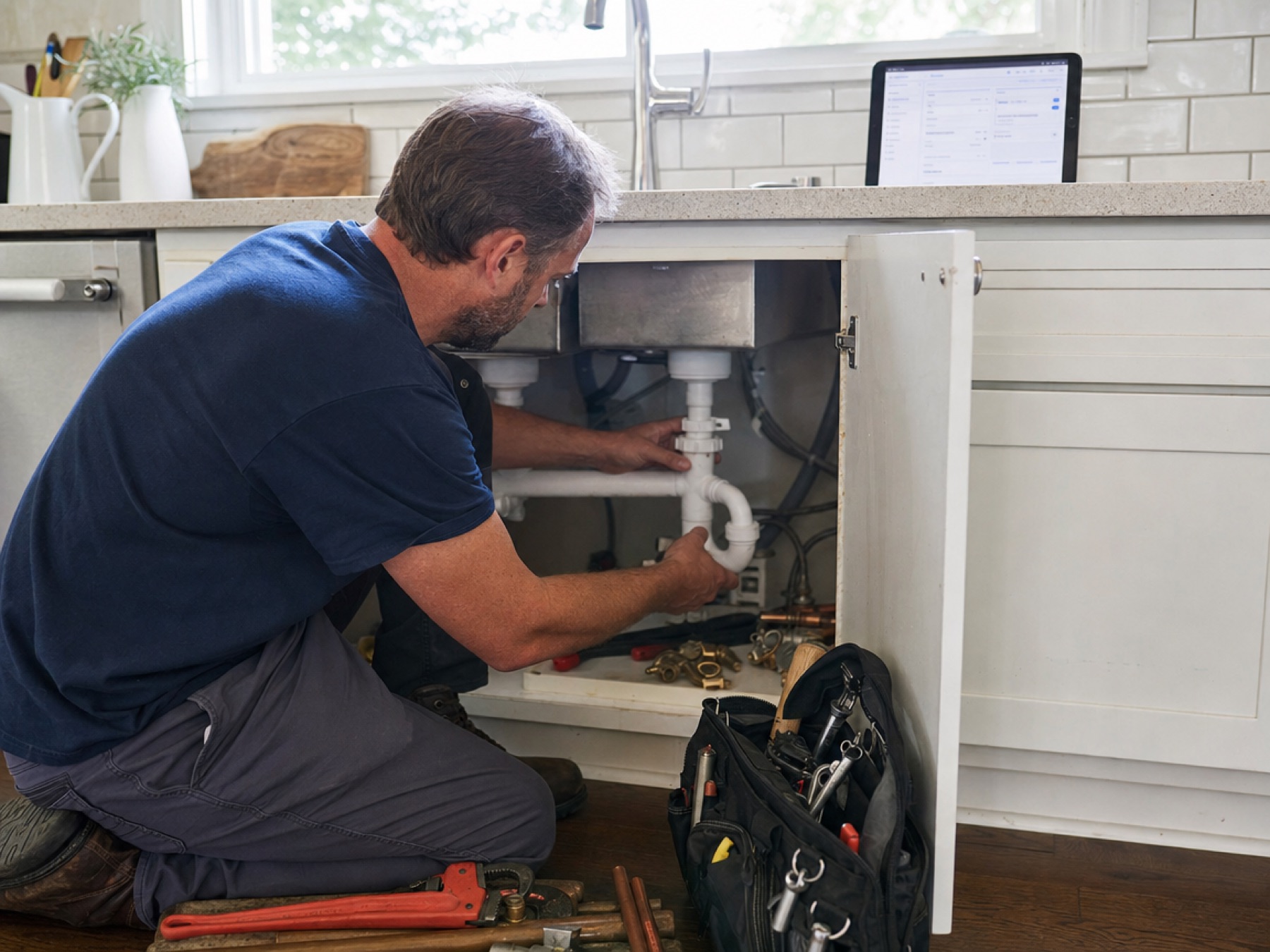 Plumber working under a kitchen sink with job details on a tablet